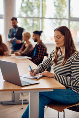 Mid adult female student takes notes while using laptop in classroom.