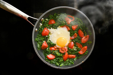 Spinach, tomatoes and fried egg in a steaming pan on a black stovetop, cooking a healthy meal for breakfast or lunch, high angle view from above, copy space