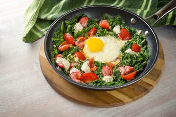 Fried egg, spinach and tomatoes with some yoghurt blobs in a frying pan on a wooden cutting board and a green kitchen towel, healthy meal, copy space, selected focus