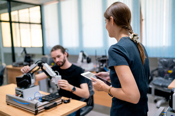 Back of Caucasian professional engineer or technician manager with casual cloth help to support and also guide staff to check small robotic machine that put on table in factory workplace.