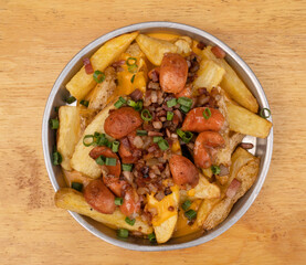 French fries. Top view of fried potatoes with sliced green onion, smoked sausages, crispy bacon and cheddar cheese sauce, in a metal dish on the wooden table.