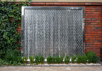 ściana wodna w ogrodzie. Metalowa ściana wodna kaskadowa. water wall in the garden, decorative water fountain cascading in garden, steel wall with a waterfall. Artificial decorative waterfall.   © kateej