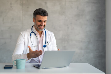Smiling confident male doctor wearing white coat and blue stethoscope sitting at desk in medical...