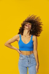 Woman with curly afro hair in a blue T-shirt on a yellow background dancing flying hair with sunglasses yellow, hand signs, look into the camera, smile with teeth and happiness, copy space