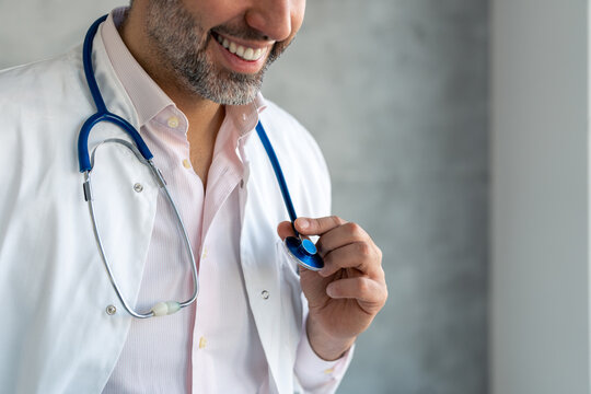Unrecognizable smiling male doctor medical expert wearing white lab coat, holding and showing blue stethoscope with one hand in medical office. Close up photo of unrecognizable handsome male doctor.