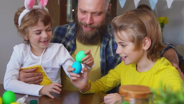 Easter Family traditions. Father and two caucasian happy children with bunny ears playing with Easter eggs with paints for holidays while sitting together at home table. Kids embrace and smile in cozy