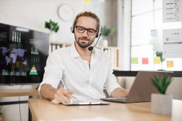 Portrait of smiling bearded young male office manager in white shirt and headset writing financial report using wireless laptop computer during working online.