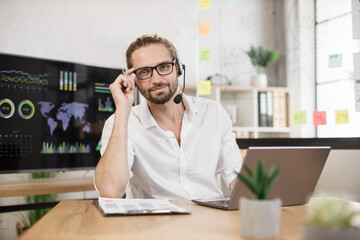 Confident businessman in stylish formal wear and headset writing notes and using modern laptop while sitting at modern office desk. Handsome company worker with beard looking at camera.