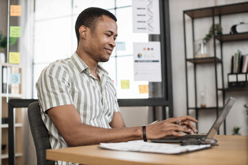 Fototapeta na wymiar Portrait of attractive young businessman sitting at office desk and working with laptop. African american guy having online video conference. Business meeting online.