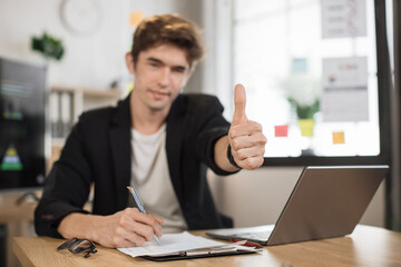 Close up portrait of young male office manager in white shirt writing financial report using wireless laptop computer during working online, looking at camera, showing thumb up