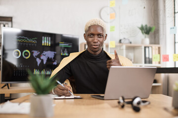Close up portrait of young african american male office manager in suit making financial report using wireless laptop computer during working online, looking at camera showing thumb up.