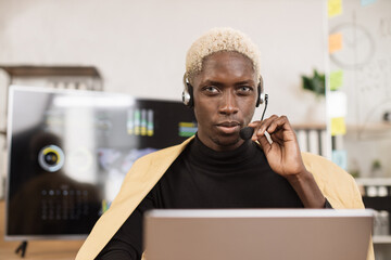 Confident african man in yellow suit using headset sitting at modern office and using wireless...