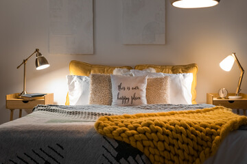 Interior of bedroom with knitted plaid on bed and glowing lamps late in evening