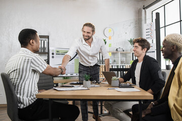 Handsome confident bearded young businessman boss shaking hand with his young african male colleague, preparing for the meeting and negotiations with multiethnic colleagues.