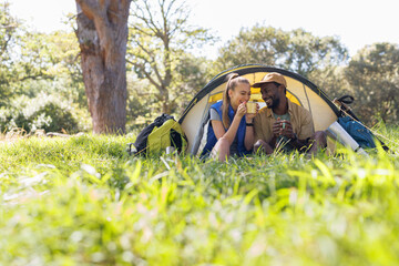 Happy, diverse couple camping in forest, sitting by tent drinking coffee and talking, copy space