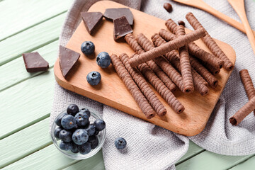 Board with delicious wafer rolls, blueberries and chocolate pieces on green wooden background