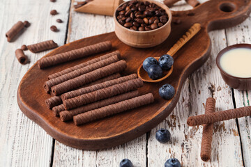 Board with delicious chocolate wafer rolls, coffee beans, condensed milk and blueberries on white wooden background