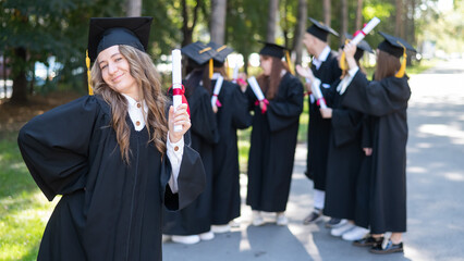 Group of happy students in graduation gowns outdoors. A young girl with a diploma in her hands in the foreground.