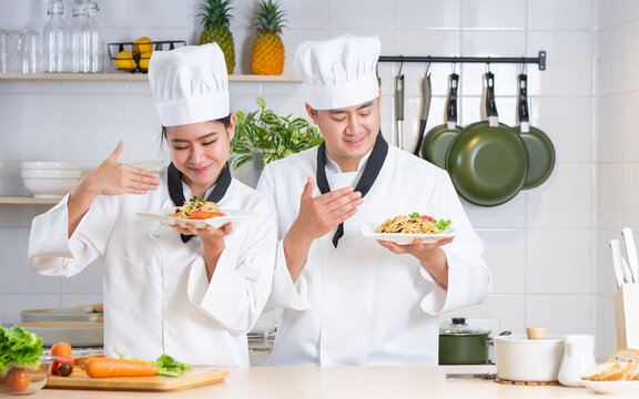 Two Asian Professional Couple Chef Wearing White Uniform, Hat, Showing Plate Of Spaghetti, Cooking In Kitchen, Making Surprising Face With Happiness. Restaurant, Food Concept.