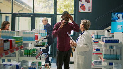Young adult entering pharmacy shop with painful headache, asking senior pharmacist about cure or medicaments. Customer suffering from migraine and rubbing temples in drugstore.