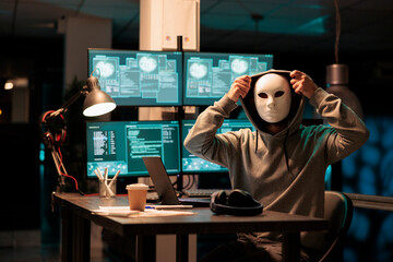 Scary masked hacker installing virus to hack system on computer, working late at night in office....