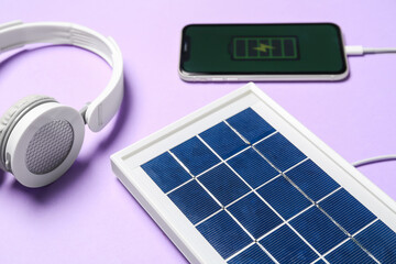 Portable solar panel charging mobile phone and headphones on lilac background, closeup