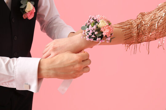 Young man tying corsage around his prom date's wrist on pink background, closeup