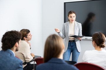 Young woman standing near interactive board and communicating with adult students during advanced...