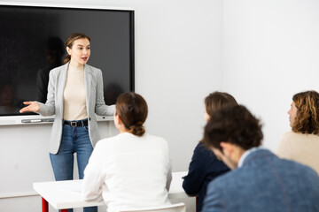 Fototapeta premium Young female business coach explaining topic while standing against group of people sitting at desks in front of her