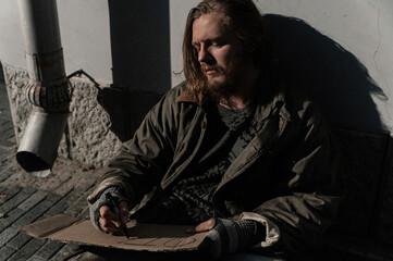 a homeless man sits on the street and writes on a sign