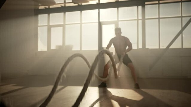 Muscular build athlete performing cardiovascular exercises, working out with battle ropes indoors
