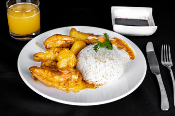 homemade chicken stew with tomatoes, onions and potatoes on a black fabric table and plate with rice, beans and orange juice