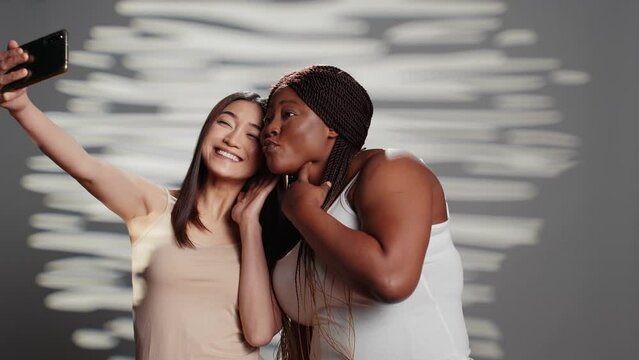 Two skincare models taking pictures on smartphone, using mobile phone to have fun with photos. Young beautiful women promoting self love and body positivity, self acceptance in studio.