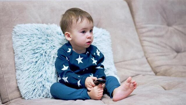 Little adorable baby mesmerized by the TV. Cute kid sits on the sofa holding remote control and watching ahead attentively.