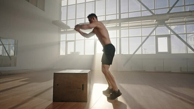 Shirtless Caucasian male athlete doing jumping squats on a wooden box, in a light smokey interior