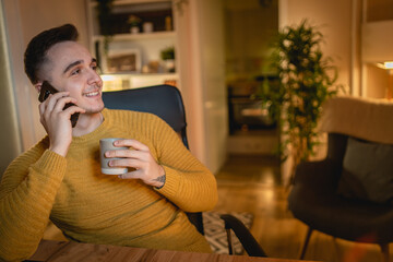 A young man is working late or studying at his home office while drinking coffee and using his mobile phone