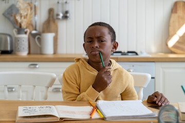 Pondering focused child boy look at camera sit at home table with pencil near mouth thinking about...
