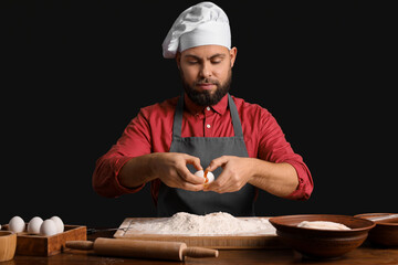 Male baker breaking egg for dough at table on dark background