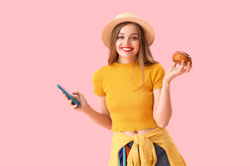 Young woman in hat with tasty bun and mobile phone on pink background