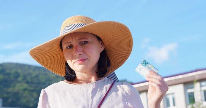 Woman straw hat against backdrop of mountains sky holds bill of money in her hands pity, looks from side to side, gets upset to tears, leaves.Compassionate sentimental tourist giving alms to homeless