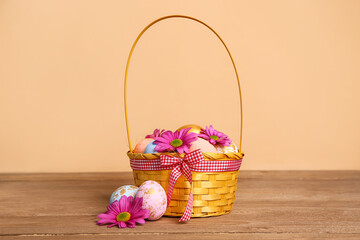 Wicker basket with painted Easter eggs and chrysanthemum flowers on wooden table near beige wall