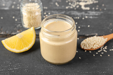 Jars with tasty tahini, sesame seeds and lemon slice on dark wooden table