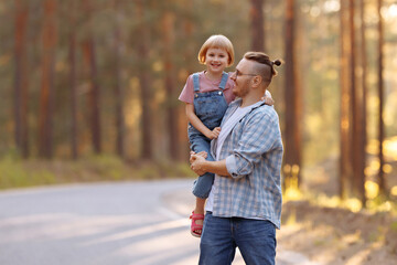 Fototapeta premium Dad holds a little daughter in his arms, they laugh merrily walking along a forest road among tall pines. Family walk in the forest at sunset, man and little girl.