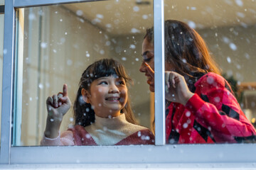Adorable child looking at the window and admire snowflakes with mother. 