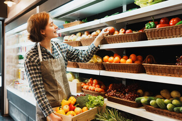 Happy Caucasian young adult employee is looking at fruit shelves and preparing to add an apple to the shelf. Saleswoman with an apron is holding an apple and managing the stock of fruit on shelves.