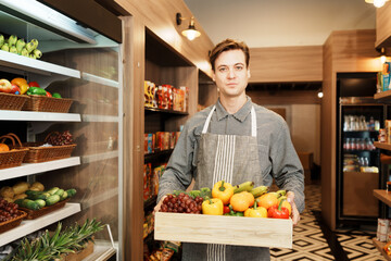 Caucasian young adult salesman is carrying the basket of fruit and preparing to add stock of fruit to the shelf. Male employee who working in the grocery store looking at the camera with confident. 