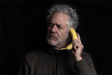 Humorous portrait of an attractive older Caucasian man with grey hair and beard, holding a banana like a telephone.  Appears confused or skeptical.