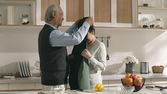 senior asian husband helping wife put on apron in kitchen at home before preparing meal