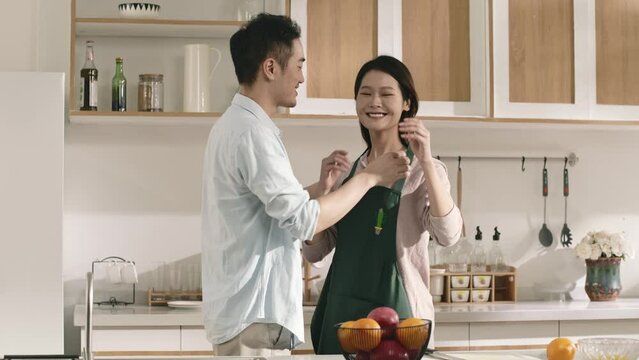 young asian husband helping wife putting on apron in kitchen at home before preparing meal