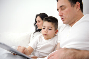 family, fatherhood, technology and people concept happy father and little son with tablet pc computer sitting on sofa at home. mom woman working in the background online on computer laptop